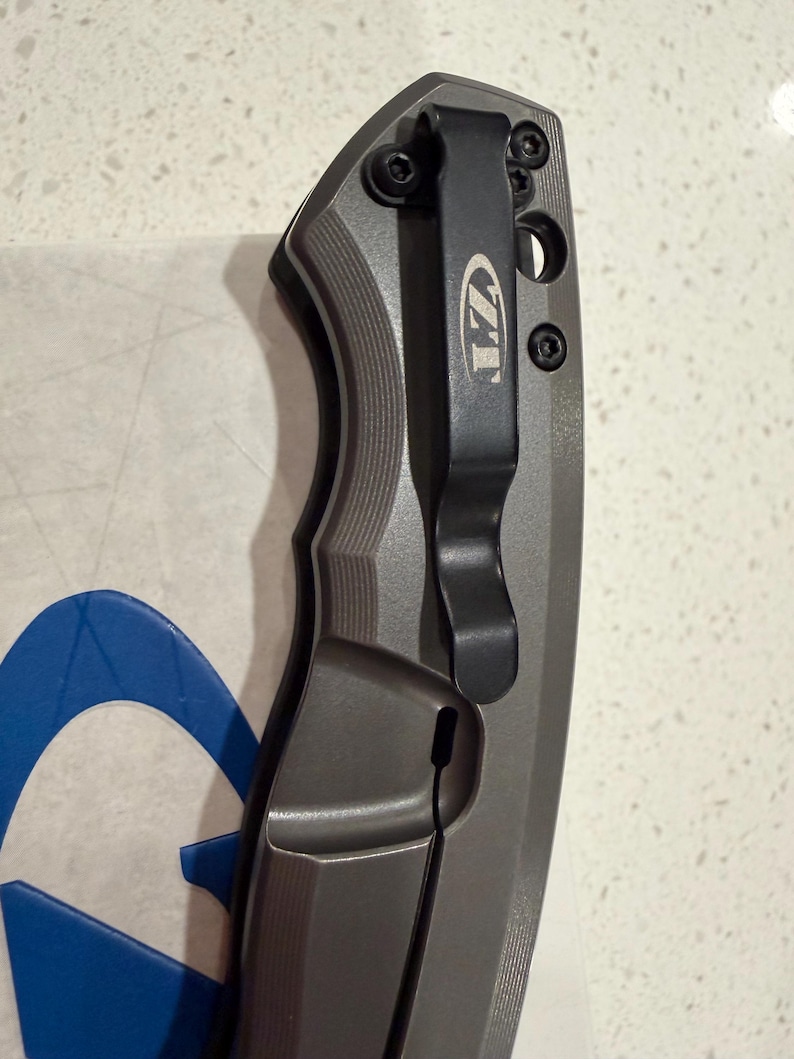 May include: A gray and black folding knife with a pocket clip. The knife has a textured handle and a black clip with the letters "ZT" engraved on it. The blade is not visible in the image.