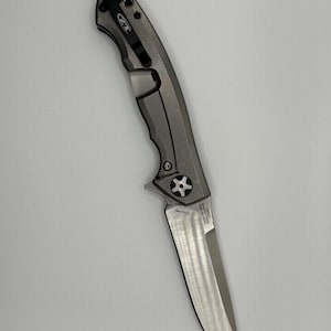 May include: A gray and silver folding knife with a partially serrated blade. The knife has a textured handle and a pocket clip. The blade is approximately 3.5 inches long. The knife is designed for everyday carry and outdoor use.