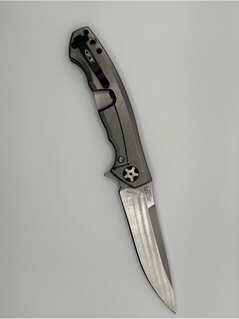 May include: A gray and silver folding knife with a partially serrated blade. The knife has a textured handle and a pocket clip. The blade is approximately 3.5 inches long. The knife is designed for everyday carry and outdoor use.