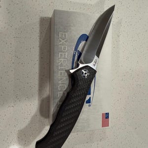 May include: A folding knife with a silver blade and a black textured handle. The knife is partially resting on a white box with the word "EXPERIENCE" printed on it. A small American flag is visible.