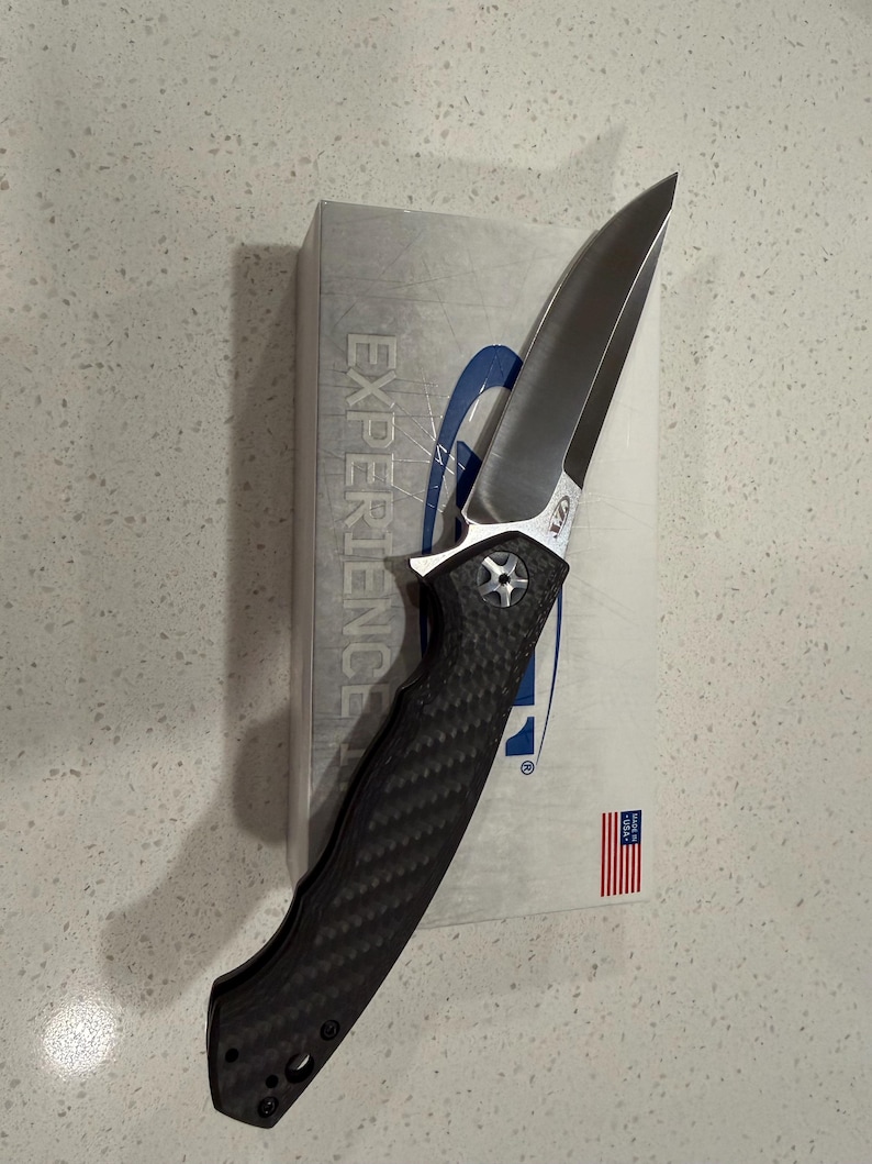 May include: A folding knife with a silver blade and a black textured handle. The knife is partially resting on a white box with the word "EXPERIENCE" printed on it. A small American flag is visible.