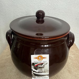 Puede incluir: Una olla de cerámica marrón oscuro con tapa y dos asas laterales. La olla tiene un acabado brillante y una etiqueta que dice "Made in Puglia - ITALY". La olla probablemente se utiliza para cocinar o almacenar.