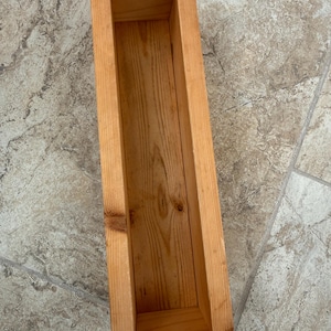 May include: A rectangular wooden planter box with a natural finish. The box is constructed from light-colored wood and features visible wood grain. The open top and rectangular shape suggest it's designed for planting flowers or herbs. The box is approximately 24 inches long.