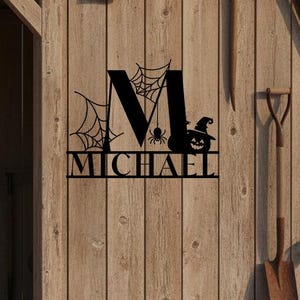 May include: A black metal wall sign featuring a large letter M decorated with spider webs, a small spider, and a jack-o'-lantern wearing a witch hat. The name MICHAEL is cut out in capital letters below the M, mounted on a wooden barn wall.