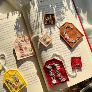 May include: A collection of handmade felt bookmarks and small decorative squares, each adorned with colorful embroidered floral designs. The pieces are in various shapes and colors, including pink, yellow, red, and brown, and are arranged on an open book.