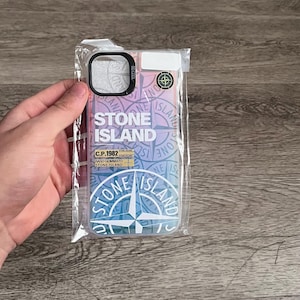 Peut inclure: Une coque de téléphone transparente avec un design dégradé coloré, avec le logo "STONE ISLAND" et les détails de la marque. La coque est dans un emballage en plastique, avec une découpe noire pour l'appareil photo.