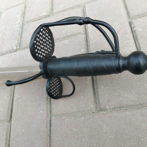 Könnte beinhalten: Ein schwarzer Rapier-Säbel mit lederummanteltem Griff und einem komplexen Parier mit perforiertem Design. Die Klinge ist silberfarben und das Gesamtdesign deutet auf eine historische oder theatralische Requisite hin.