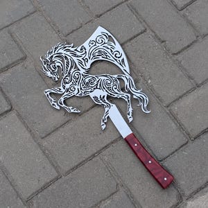 Puede incluir: Un hacha de metal con una hoja plateada recortada en forma de caballo con intrincados patrones de remolinos. El hacha tiene un mango recto y liso con un acabado pulido de color rojo intenso y dos pequeños remaches plateados en la empuñadura.