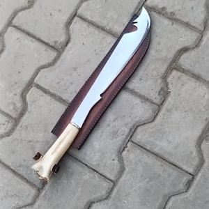 May include: A large, polished steel machete with a curved blade, set in a brown leather sheath. The handle is light-colored wood with a brass bolster. The machete is designed for heavy-duty cutting tasks.