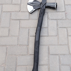 May include: A black and silver axe with a curved, black handle. The axe head features a silver blade with black and silver accents. The handle is wrapped in black tape near the head. The axe is lying on a gray brick surface.