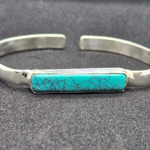 May include: A silver cuff bracelet featuring a rectangular, horizontally oriented turquoise stone inlay. The stone is bright teal with a dark, intricate web-like matrix pattern. The metal band has a smooth, polished finish and an open back design.