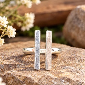 Peut inclure: Une bague en argent composée de deux barres rectangulaires verticales parallèles fixées sur un anneau lisse. Les deux barres présentent une finition texturée et mate qui contraste avec la surface polie et réfléchissante de l'anneau.