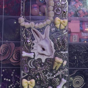 Op de afbeelding: Transparante telefoonhoes met een decoratief ontwerp. De hoes heeft een witte konijnenillustratie, samen met verschillende bedels, waaronder tanden, strikken, een spin, een kruis en dobbelstenen. De hoes is versierd met strass.