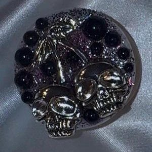 Op de afbeelding: Een rond, zilverkleurig accessoire met een schedelthema, met een glitterende paarse en zilveren achtergrond. Het ontwerp bevat twee schedels en verschillende zwarte, bolvormige versieringen. Het accessoire heeft een gotische of alternatieve uitstraling.