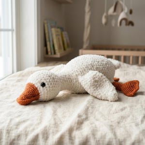 Könnte beinhalten: Ein weißes, gehäkeltes Gänse-Stofftier mit einer strukturierten, weichen Garnoberfläche. Die Gans hat einen leuchtend orangefarbenen, gehäkelten Schnabel und passende orangefarbene Schwimmfüße. Sie hat ein einzelnes schwarzes Perlenauge.