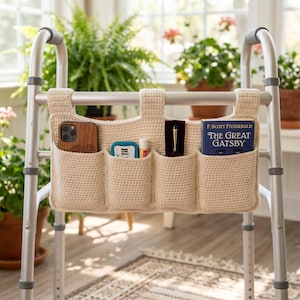 May include: A beige crochet walker bag with four pockets attached to a silver metal walker. The pockets contain a wooden-cased smartphone, a small tin, a lip balm, a black pen case, and a copy of the book The Great Gatsby by F. Scott Fitzgerald.