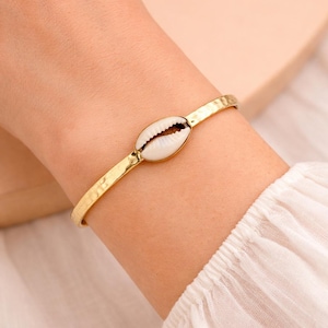 May include: Gold-coloured bracelet with a white and brown seashell centrepiece. The bracelet has a hammered texture and is worn on a wrist with a white long-sleeved shirt. A simple, elegant piece of jewellery.