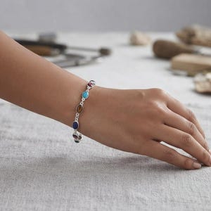 May include: A silver chain bracelet featuring a series of small, oval-shaped gemstones in various colors, including blue, brown, turquoise, and dark red, each set in a polished silver bezel connected by delicate silver links.