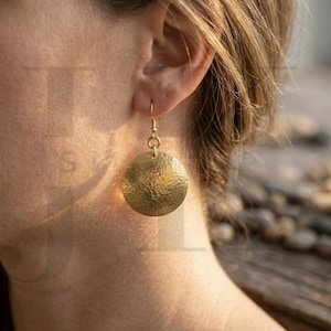 May include: A close-up of a gold-colored, hammered-metal disc earring. The earring is round and hangs from a small gold hook. The texture of the metal is visible, and the earring is catching the light.
