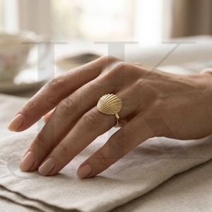 Op de afbeelding: Een goudkleurige ring met een schelpmotief. De ring heeft een ronde, geribbelde schelpvorm en een dunne band. De ring wordt gedragen aan een hand met neutrale nagellak, rustend op een beige stof.