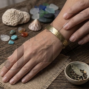 May include: A hammered gold-tone cuff bracelet on a wrist. The bracelet has a textured surface. The background includes a small bowl with herbs, seashells, and colorful stones on a wooden surface.