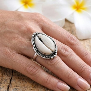 May include: A silver ring with a large, oval white seashell. The ring has a detailed, textured border. The ring is set against a natural wood background. A statement piece of jewellery.