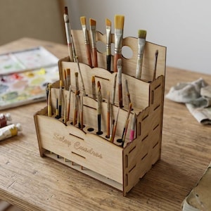 Puede incluir: Un organizador de pinceles de madera con múltiples compartimentos, lleno de varios pinceles. El organizador está sobre una mesa de madera, junto a una paleta, tubos de pintura y un paño. El texto "Solay Creations" es visible.