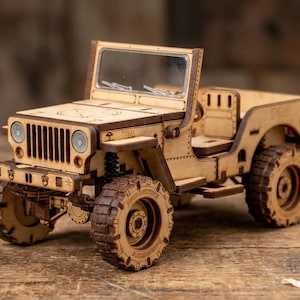 Può includere: Modello in legno di una Jeep d'epoca, con dettagli intagliati al laser. Il modello è di colore beige, con pneumatici visibili, parabrezza e una griglia anteriore dettagliata. La Jeep è esposta su una superficie di legno, con trucioli di legno sparsi.