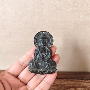Può includere: Un piccolo ciondolo a forma di Buddha di colore scuro. Il ciondolo raffigura una figura di Buddha seduta con le mani in posa meditativa, circondata da un'aureola circolare. Il ciondolo ha un foro nella parte superiore per appenderlo.