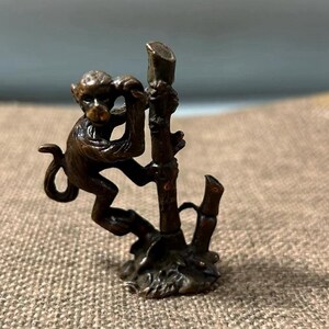 Könnte beinhalten: Eine bronzefarbene Affenfigur, die einen Ast hinaufklettert. Der Affe ist detailliert mit sichtbaren Gesichtszügen und Fellstruktur. Der Ast hat eine raue, strukturierte Oberfläche. Die Figur ist wahrscheinlich ein Dekorationsartikel oder Sammlerstück.