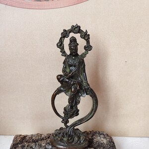 Può includere: Statua color bronzo di una figura seduta con un'aureola, che tiene una figura più piccola. La statua è montata su una base decorativa e posta su uno sfondo neutro. Il design della statua include dettagli intricati.