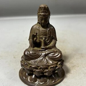 Può includere: Una statua color bronzo di una figura seduta, forse una divinità, con lineamenti sereni e abiti ornati. La figura è seduta su una base a forma di fiore di loto, con le mani in un gesto di benedizione o insegnamento.