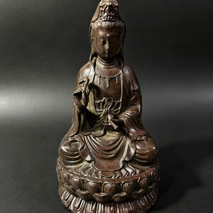 Può includere: Statua color bronzo di una figura seduta, forse una divinità, con lineamenti sereni e copricapo ornato. La figura è seduta su una base a forma di fiore di loto, con le mani in un gesto di benedizione o insegnamento. La statua ha un design dettagliato.