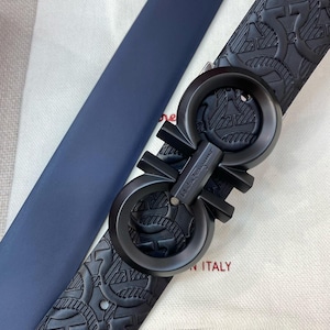 May include: A close-up of a black leather belt with a textured pattern and a large, black buckle. The belt is navy blue on the reverse side. The buckle has the brand name on it. The words "ITALY" are visible.