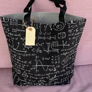 May include: A black tote bag featuring a white mathematical formula print, including equations, graphs, and geometric shapes. The bag has two black fabric handles and a light gray interior lining. A blank beige rectangular tag hangs from one handle.