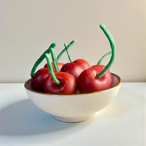 Puede incluir: Un cuenco de cerámica blanco lleno de varias cerezas rojas con tallos verdes. El cuenco tiene un borde marrón y está sobre una superficie blanca. Las cerezas son el foco, su color contrasta con el cuenco.