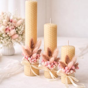 May include: Three cylindrical beeswax candles with a honeycomb texture, each decorated with a cluster of dried pink and brown bunny tail grass and small pink flowers, secured with a light cream-colored satin ribbon tied in a bow at the base.