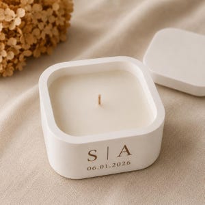 May include: A square white candle in a matte container with a single centred wick. The front of the container features the printed text S | A above the date 06.01.2026 in a dark brown serif font. A matching square white lid sits to the side.