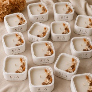 May include: A collection of square white candles, each featuring a single wick and a cluster of dried flowers in shades of beige and brown. The front of each candle container is printed with the letters S | A and the date 06.01.2026 in dark text.