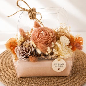 May include: A clear plastic gift bag containing a floral arrangement of dried flowers in shades of peach, cream, and brown. The base is wrapped in light brown paper with a round tag that reads HANDMADE with love and a small heart icon.