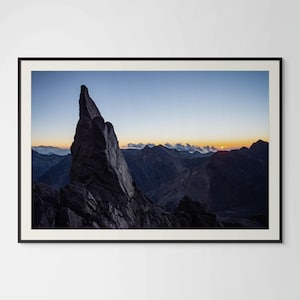 Puede incluir: Fotografía enmarcada de un paisaje montañoso al amanecer. Una oscura y dentada formación rocosa domina el primer plano, contrastando con el cielo azul y naranja, creando una escena serena y majestuosa.