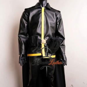 Puede incluir: Un conjunto de chaqueta y pantalón de cuero negro con una franja vertical amarilla en el centro de la chaqueta, un cinturón utilitario negro con una hebilla dorada y una capa negra. La prenda se muestra en un maniquí negro.
