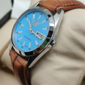 May include: A silver-toned Seiko 5 automatic watch with a light blue dial and brown leather strap. The watch face displays the day and date. The watch is presented on a cream-colored cushion.