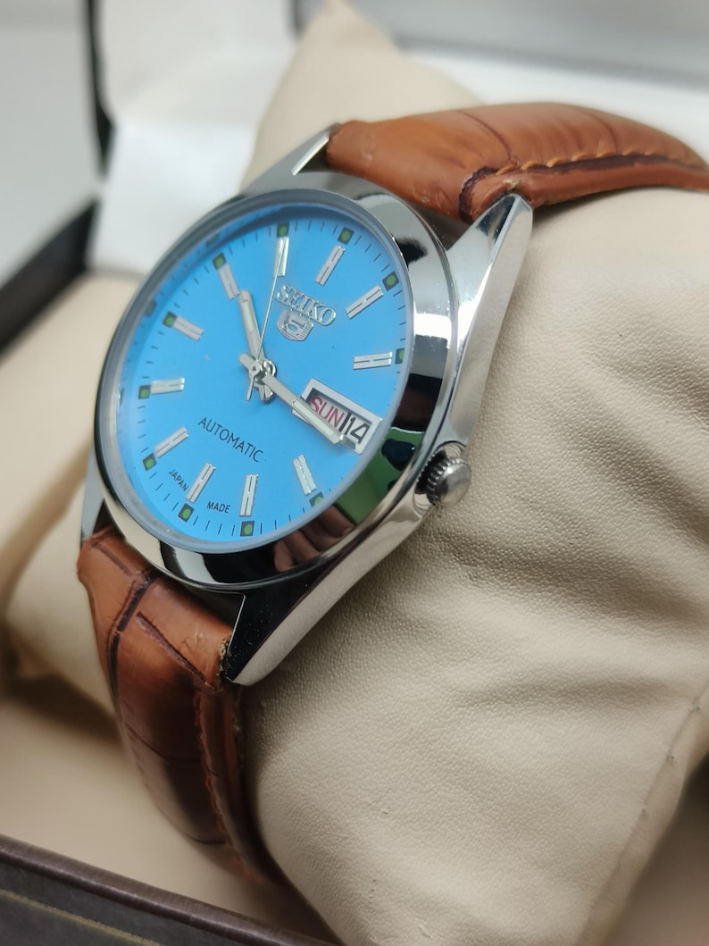 May include: A silver-toned Seiko 5 automatic watch with a light blue dial and brown leather strap. The watch face displays the day and date. The watch is presented on a cream-colored cushion.