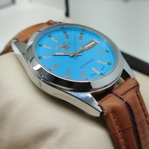 May include: A silver-toned Seiko automatic watch with a light blue face and brown leather band. The watch face features the Seiko logo, day and date indicators, and the word "AUTOMATIC".