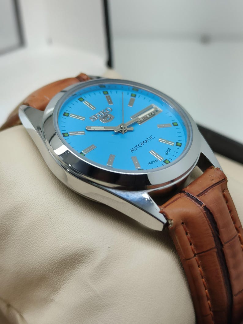 May include: A silver-toned Seiko automatic watch with a light blue face and brown leather band. The watch face features the Seiko logo, day and date indicators, and the word "AUTOMATIC".