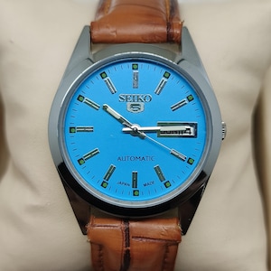 May include: A Seiko 5 automatic watch with a light blue dial and a brown leather strap. The watch face features silver hands and markers, with the day and date displayed at the 3 o'clock position. The watch is marked "Japan Made".