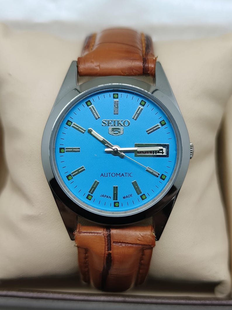 May include: A Seiko 5 automatic watch with a light blue dial and a brown leather strap. The watch face features silver hands and markers, with the day and date displayed at the 3 o'clock position. The watch is marked "Japan Made".