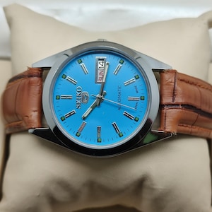 May include: A Seiko automatic watch with a light blue face and a brown leather band. The watch has a silver-colored bezel and displays the day and date. The watch is presented on a cream-colored pillow.