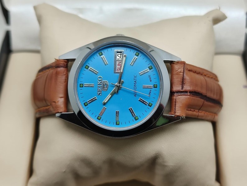 May include: A Seiko automatic watch with a light blue face and a brown leather band. The watch has a silver-colored bezel and displays the day and date. The watch is presented on a cream-colored pillow.